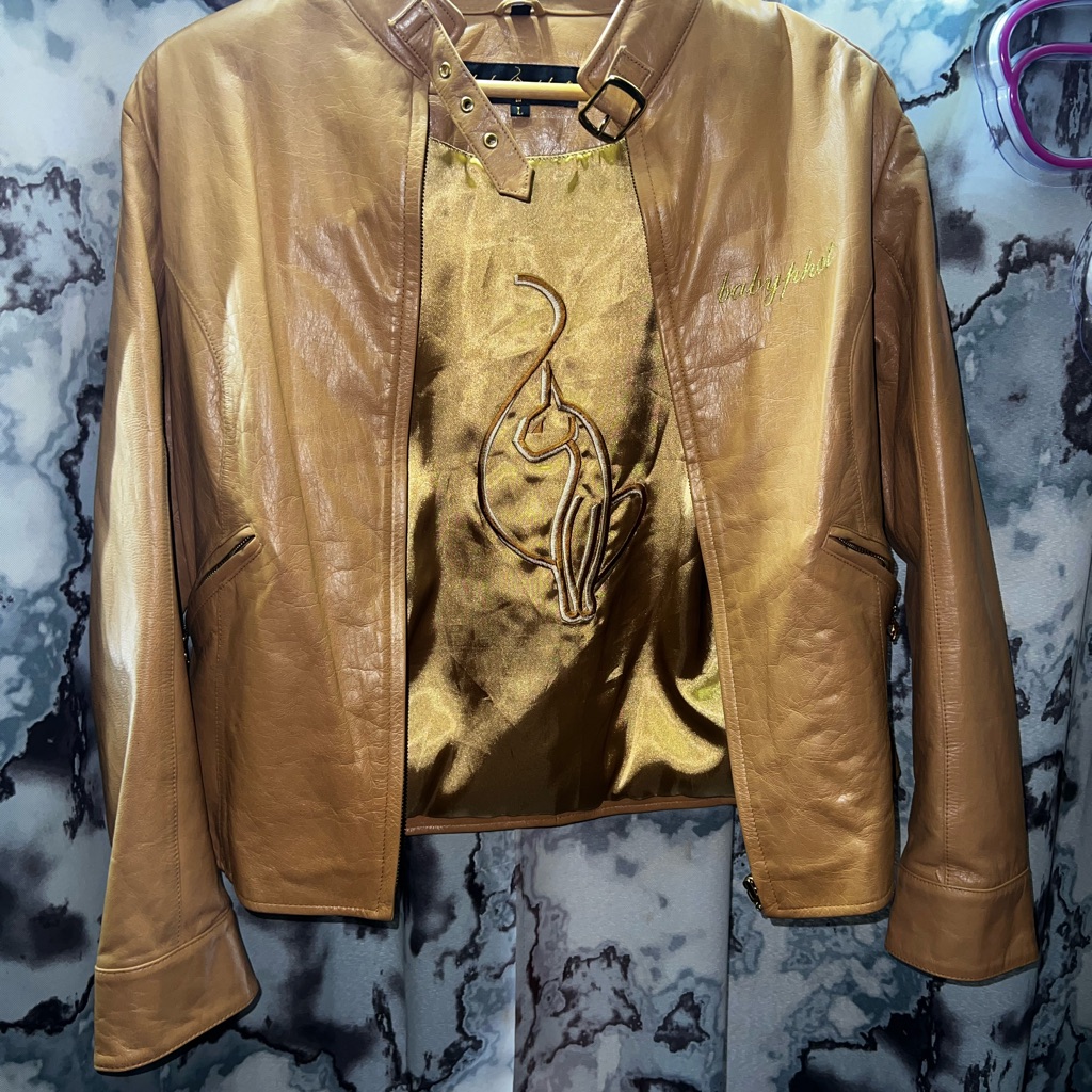 Vintage Baby Phat gold leather jacket with the cat logo embroidered on the lining, shown on a hanger
